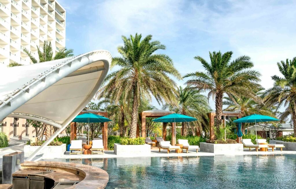 Luxury hotel outdoor pool area with palm trees, teak wood furniture, sun loungers, teal umbrellas, and modern cabanas.